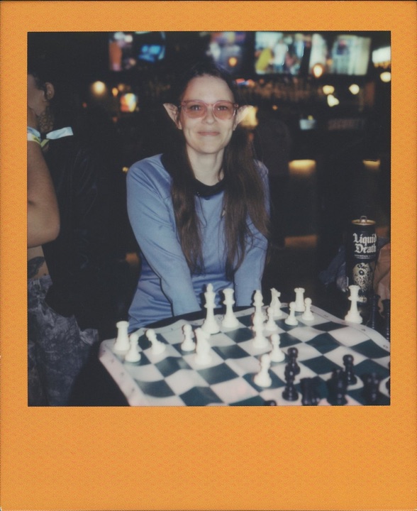 Polaroid of Birdie in elf ears, smiling behind a chessboard